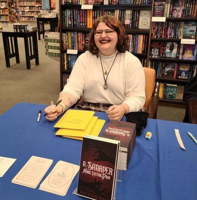An image of Alex tabling at a book event. They are wearing a white turtleneck and a brown plaid skirt and holding a pen to sign books with. Before them is a stack of books on a table.